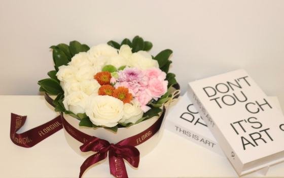 Arranjo floral em caixa redonda bege, composto por rosas brancas, flores em tons de rosa, laranja e verde, emolduradas por folhagens verdes naturais. A caixa é decorada com fita bordô personalizada da Florgiul e está posicionada sobre uma superfície branca ao lado de dois livros decorativos.