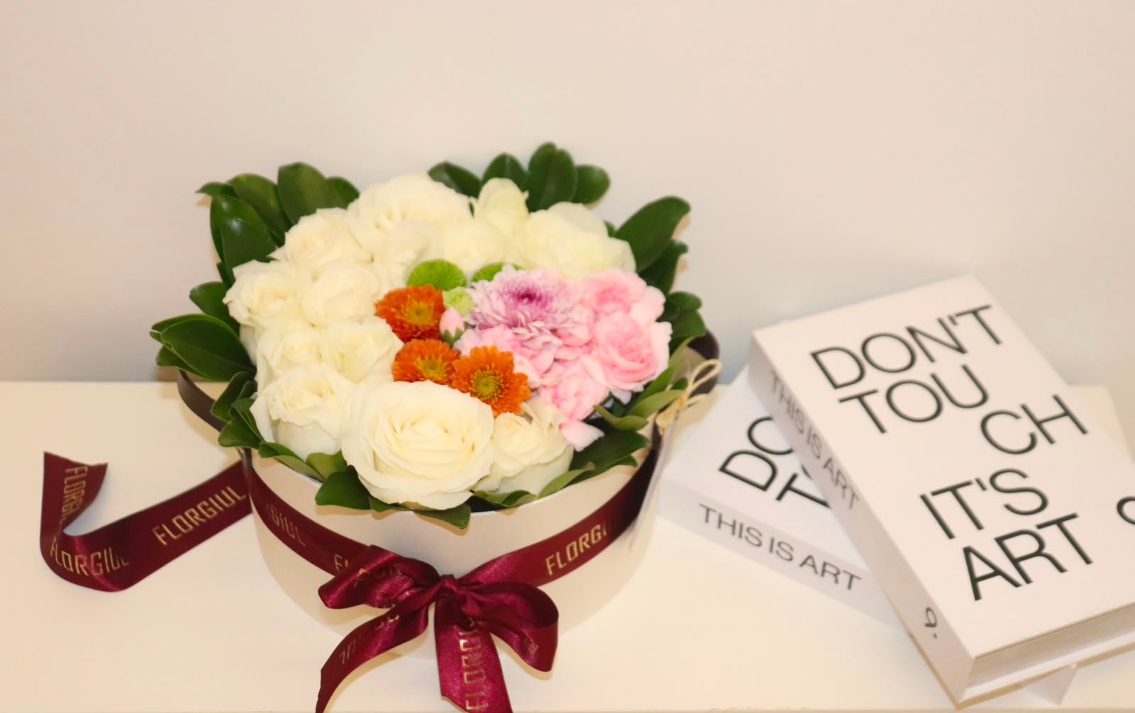 Arranjo floral em caixa redonda bege, composto por rosas brancas, flores em tons de rosa, laranja e verde, emolduradas por folhagens verdes naturais. A caixa é decorada com fita bordô personalizada da Florgiul e está posicionada sobre uma superfície branca ao lado de dois livros decorativos.