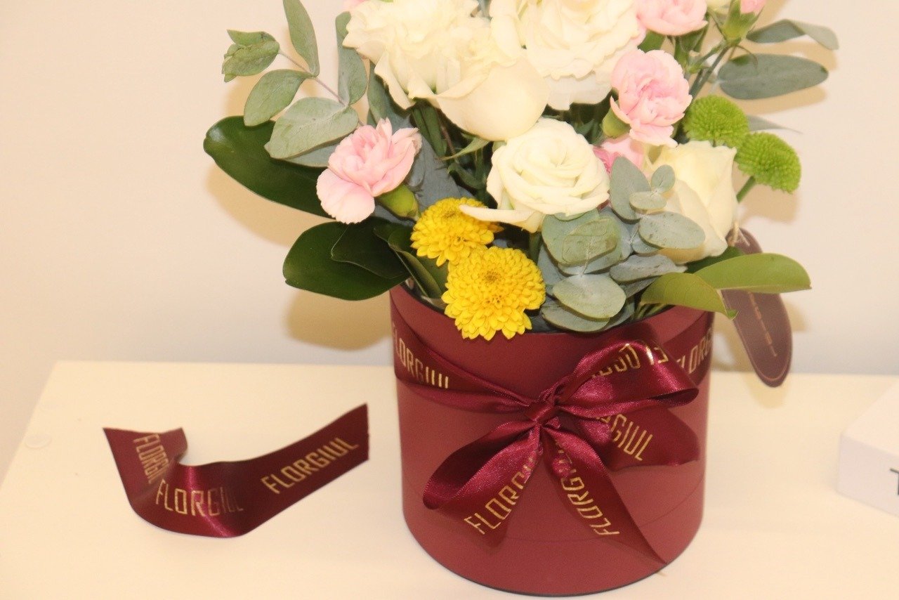 Arranjo floral em caixa redonda bordô, composto por rosas brancas, mini cravos cor-de-rosa, flores amarelas e verdes, folhagens naturais e eucalipto. A caixa é decorada com fita personalizada da Florgiul e está posicionada sobre uma superfície branca ao lado de dois livros decorativos. — foto 2
