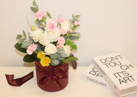 Arranjo floral em caixa redonda bordô, composto por rosas brancas, mini cravos cor-de-rosa, flores amarelas e verdes, folhagens naturais e eucalipto. A caixa é decorada com fita personalizada da Florgiul e está posicionada sobre uma superfície branca ao lado de dois livros decorativos.