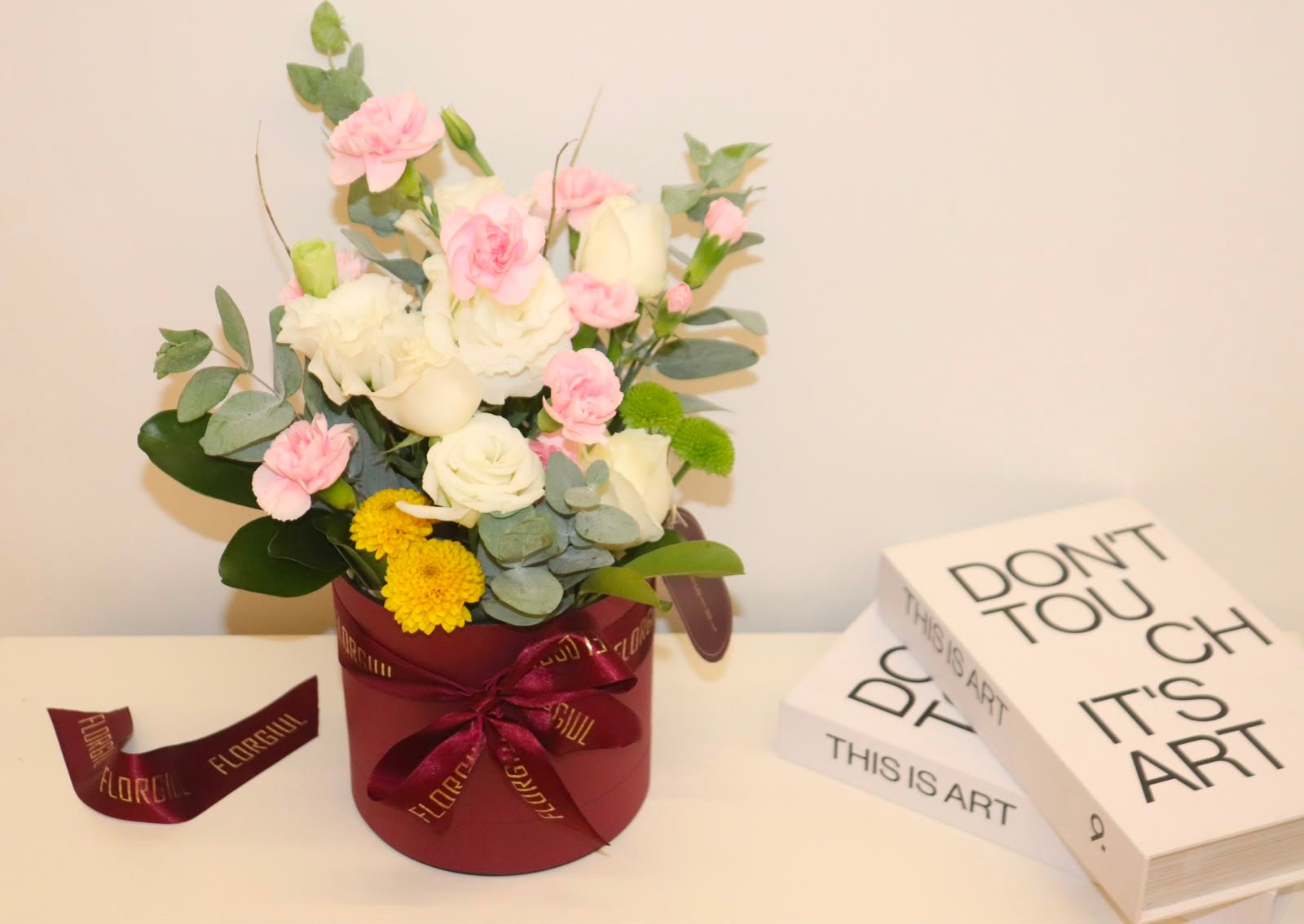 Arranjo floral em caixa redonda bordô, composto por rosas brancas, mini cravos cor-de-rosa, flores amarelas e verdes, folhagens naturais e eucalipto. A caixa é decorada com fita personalizada da Florgiul e está posicionada sobre uma superfície branca ao lado de dois livros decorativos.
