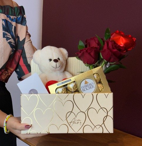 Caixa presenteável contendo um arranjo com três rosas colombianas vermelhas e folhagens naturais em vaso de vidro, acompanhadas por um ursinho de pelúcia branco com coração vermelho, uma caixa de chocolates Ferrero Rocher e um balão vermelho em formato de coração. A composição está organizada em uma caixa decorativa sobre fundo neutro. — foto 2