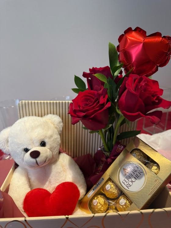 Caixa presenteável contendo um arranjo com três rosas colombianas vermelhas e folhagens naturais em vaso de vidro, acompanhadas por um ursinho de pelúcia branco com coração vermelho, uma caixa de chocolates Ferrero Rocher e um balão vermelho em formato de coração. A composição está organizada em uma caixa decorativa sobre fundo neutro.