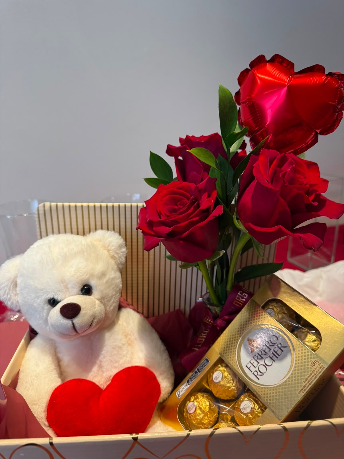 Caixa presenteável contendo um arranjo com três rosas colombianas vermelhas e folhagens naturais em vaso de vidro, acompanhadas por um ursinho de pelúcia branco com coração vermelho, uma caixa de chocolates Ferrero Rocher e um balão vermelho em formato de coração. A composição está organizada em uma caixa decorativa sobre fundo neutro.