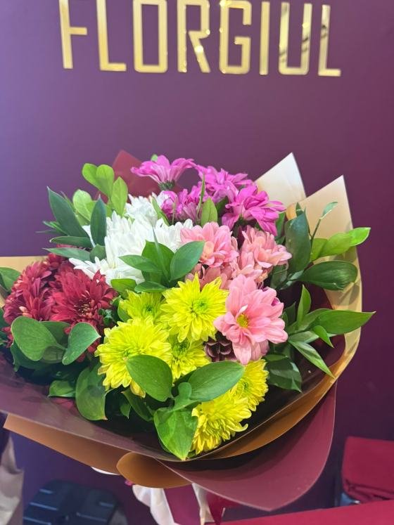 Bouquet composto por flores do campo em tons de branco, rosa, amarelo, vinho e lilás, harmonizadas com folhagens verdes naturais. O arranjo é envolto em papéis decorativos em tons de bordô e dourado, formando uma composição colorida e delicada, fotografada em frente à identidade visual da Florgiul.