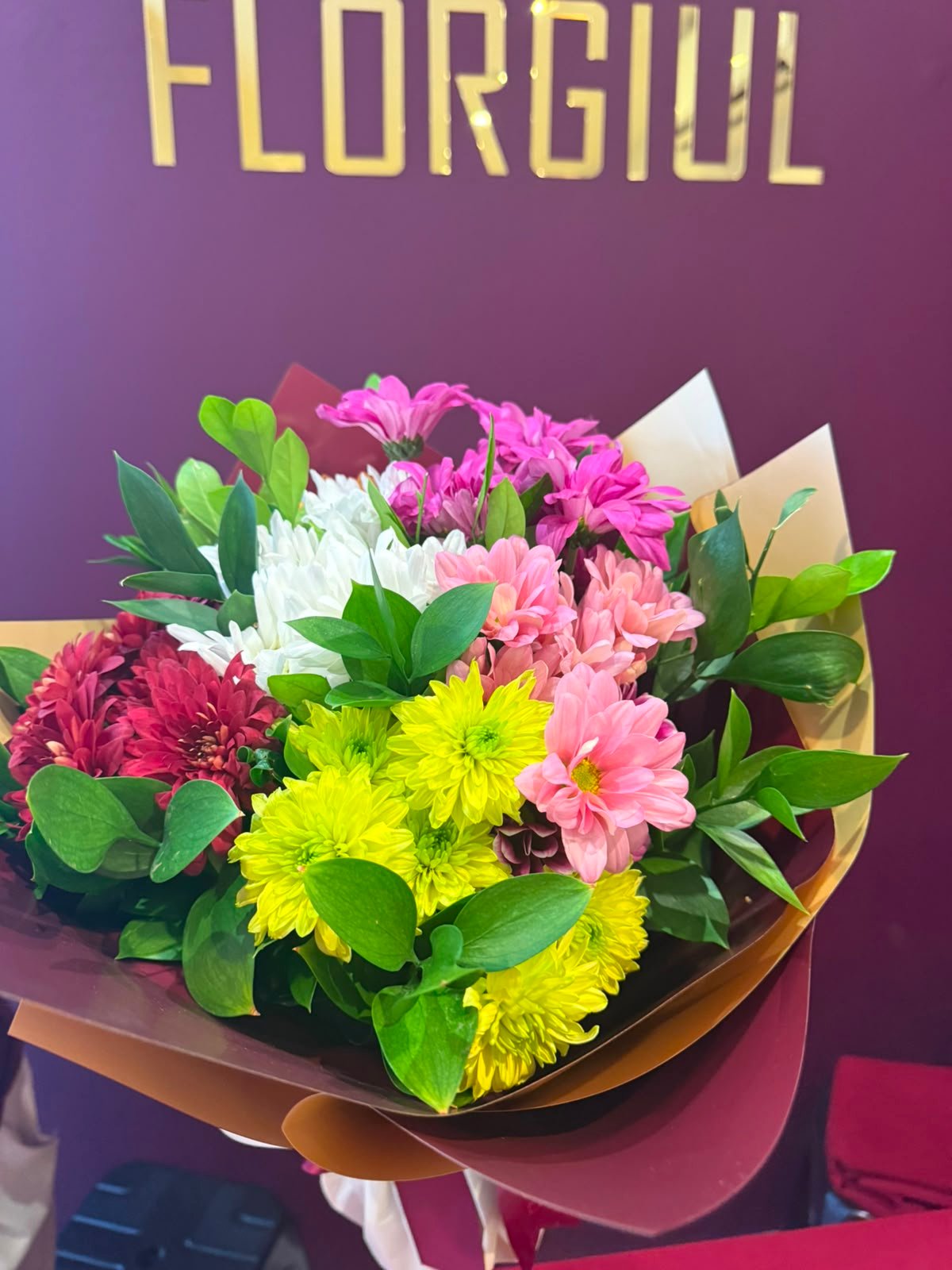 Bouquet composto por flores do campo em tons de branco, rosa, amarelo, vinho e lilás, harmonizadas com folhagens verdes naturais. O arranjo é envolto em papéis decorativos em tons de bordô e dourado, formando uma composição colorida e delicada, fotografada em frente à identidade visual da Florgiul.