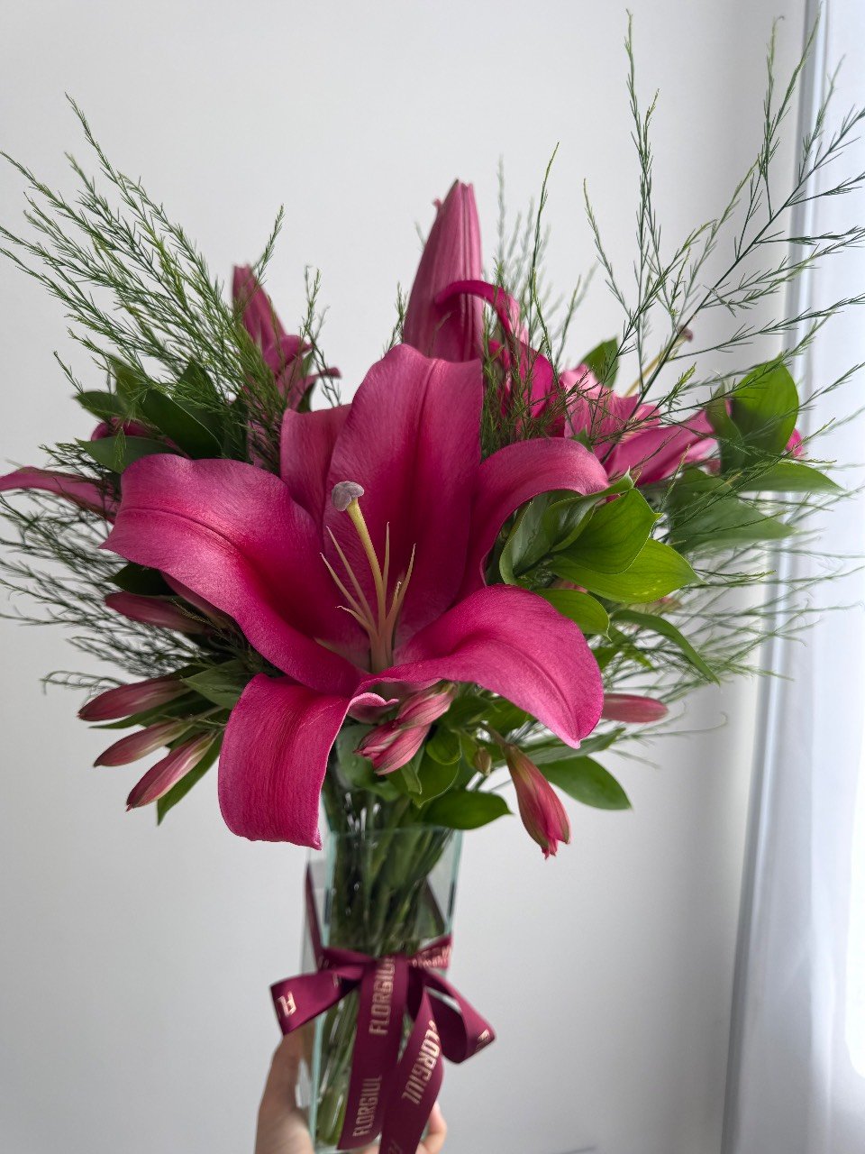 Sacola bordô personalizada da Florgiul contendo um pequeno arranjo floral com flores da semana e folhagens verdes. À frente da sacola há um mimo embalado com fita personalizada da marca. — foto 3