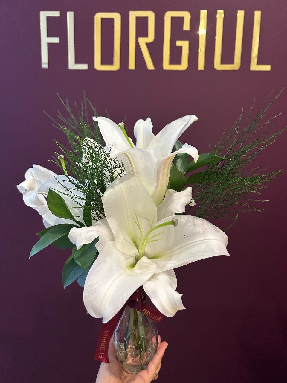Sacola bordô personalizada da Florgiul contendo um pequeno arranjo floral com flores da semana e folhagens verdes. À frente da sacola há um mimo embalado com fita personalizada da marca. — foto 4