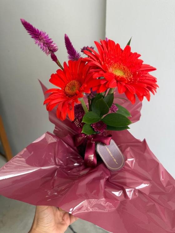 Sacola bordô personalizada da Florgiul contendo um pequeno arranjo floral com flores da semana e folhagens verdes. À frente da sacola há um mimo embalado com fita personalizada da marca.