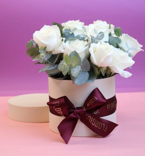 Flower Box redonda em tom claro, personalizada com o logotipo Florgiul e laço de cetim. A composição reúne 12 rosas brancas naturais cercadas por folhagens de eucalipto, formando um arranjo clássico e elegante.