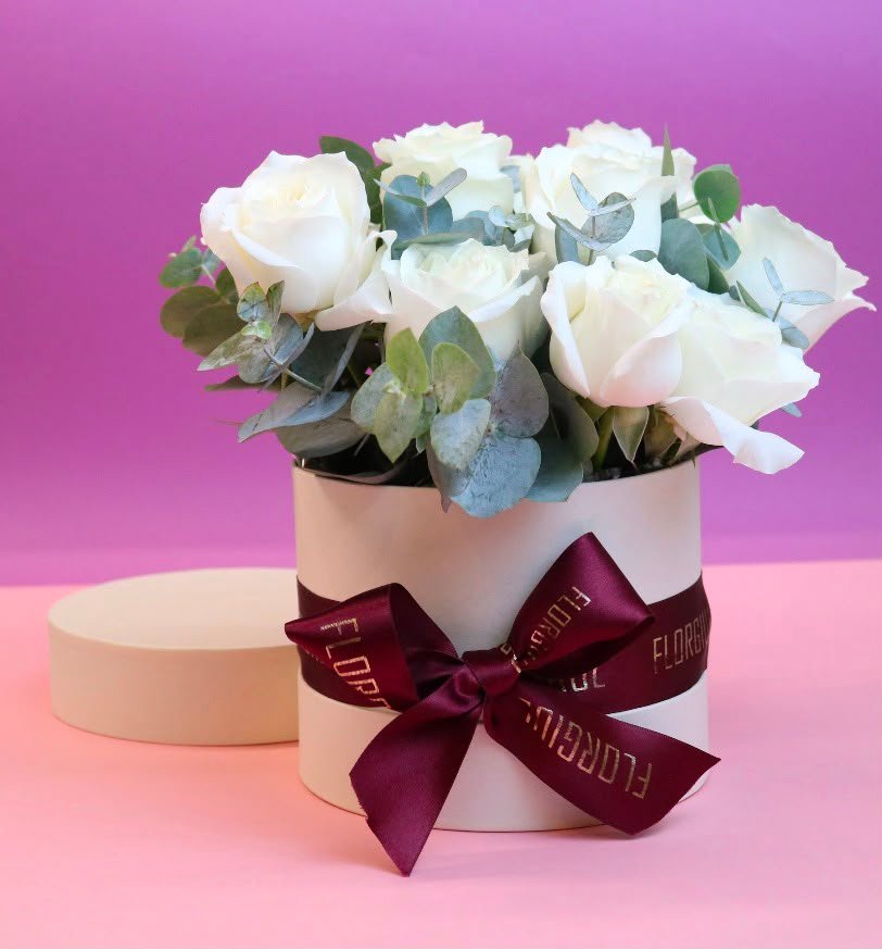 Flower Box redonda em tom claro, personalizada com o logotipo Florgiul e laço de cetim. A composição reúne 12 rosas brancas naturais cercadas por folhagens de eucalipto, formando um arranjo clássico e elegante.